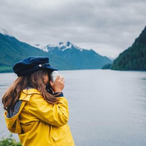 Photo of a girl standing on the shore taking photos of mountains in the distance.