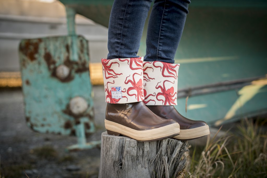 Photo of person wearing brown XTRATUF boots with the tops rolled down, featuring a red Octopus Salmon Sisters design. Person is standing on top of tree stump in front of an old, grounded fishing boat