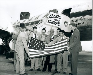 A photo of a group of men standing outside of an Alaska Airlines DC-4 aircraft with a 48-star American flag. One man in the middle is placing a 49th star on the flag.