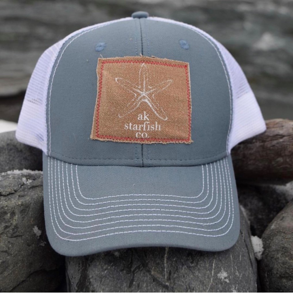 This is a photo of a trucker hat sitting on rocks on a beach. The hat features a Carhartt material patch with the akstarfish co flagship design.