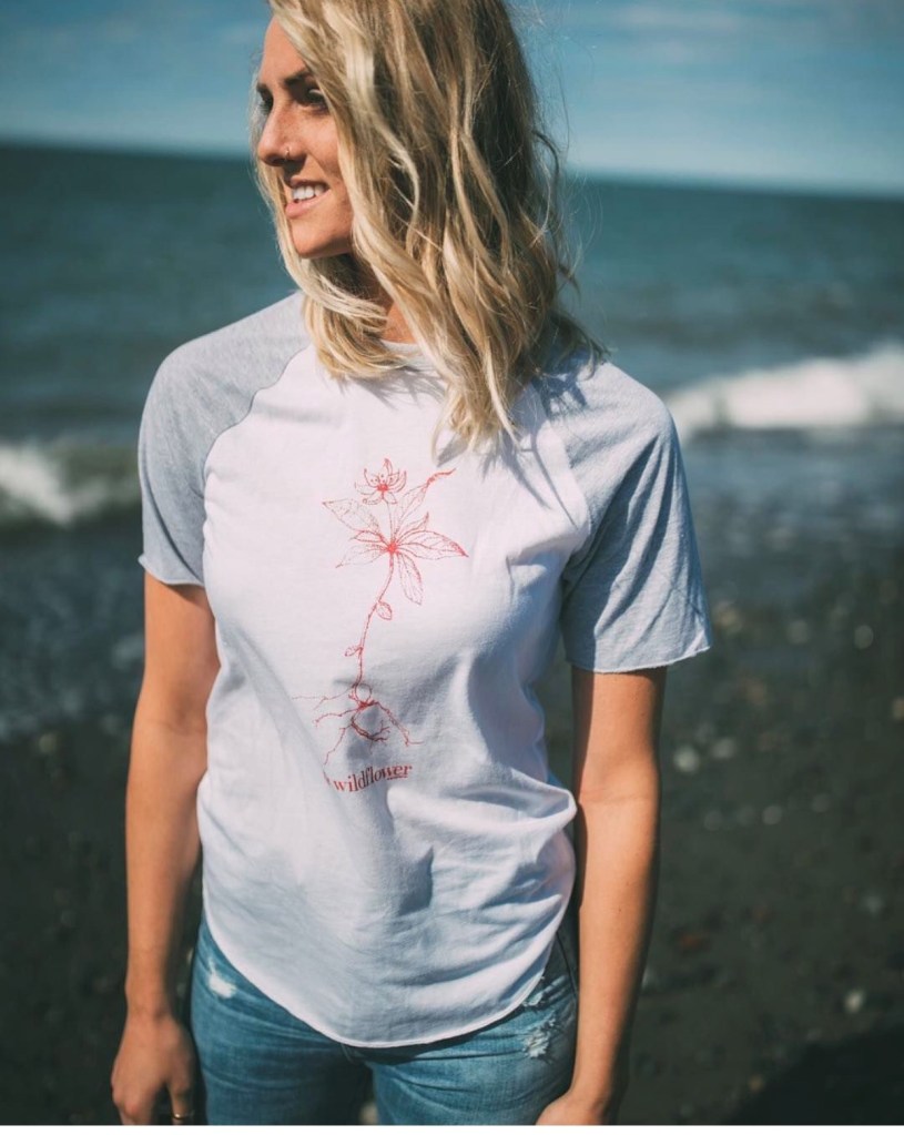 This is a photo of a woman standing on a beach wearing an AK Wildflower branded t-shirt from akstarfishco.