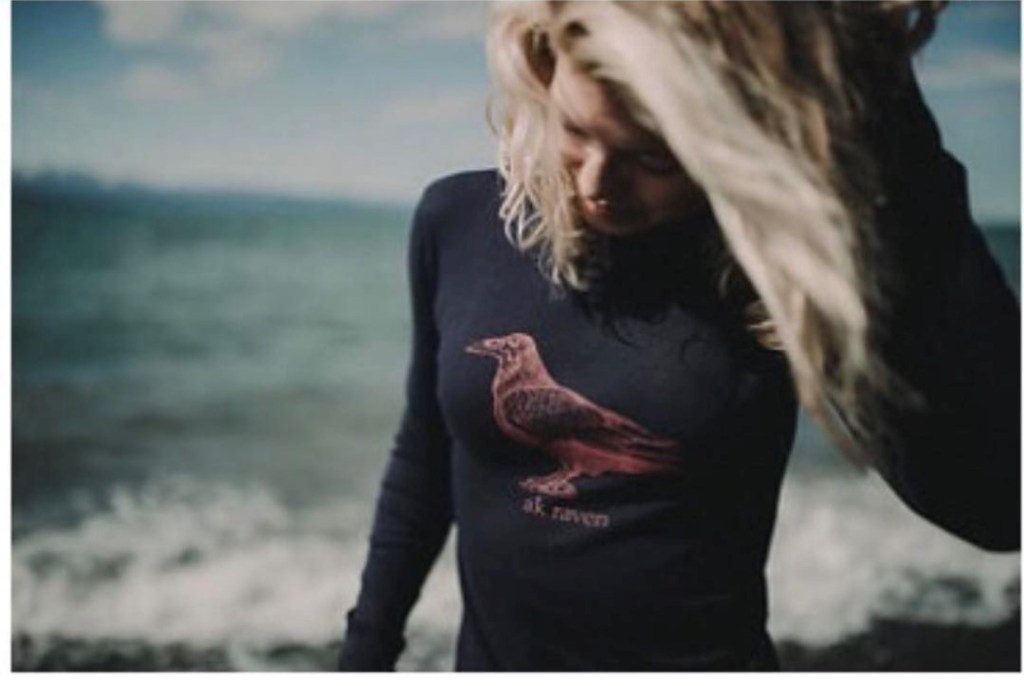 This is a photo of a woman on a beach wearing a black sweatshirt with a red AK raven design from akstarfishco
