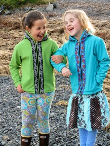 This is a photo of two young girls walking arm in arm on a beach. The girl on the left is wearing a green softshell jacket and the girl on the right is wearing a blue softshell jacket.