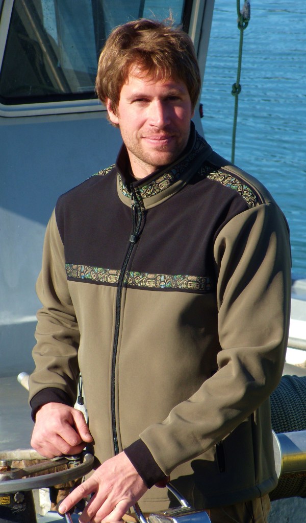 This is a photo of a man wearing a brown jacket with an Alaska Native trim , while on a fishing boat.
