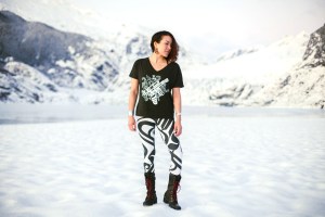 This is a photo of a woman standing on a snowy beach in front of a glacier wearing a t-shirt and leggings featuring contemporary Alaska Native artwork by Trickster Co.