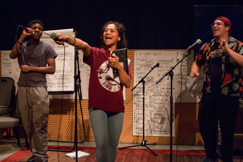 This is a photo of three students on a practice stage singing/rapping. The student in the middle is holding her microphone to the audience asking them to sing along.