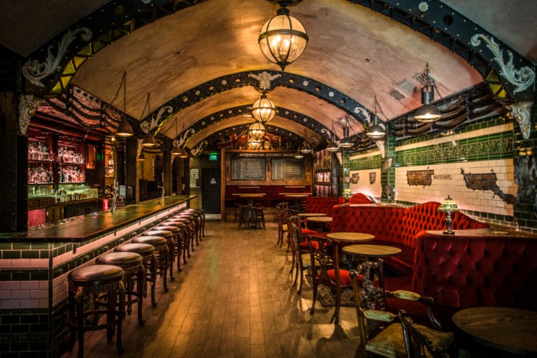 This is a photo of the Whitechapel Bar. It features wooden floors, a bar on the left with tiling and red sofas on the right. The ceiling is dome-shaped.